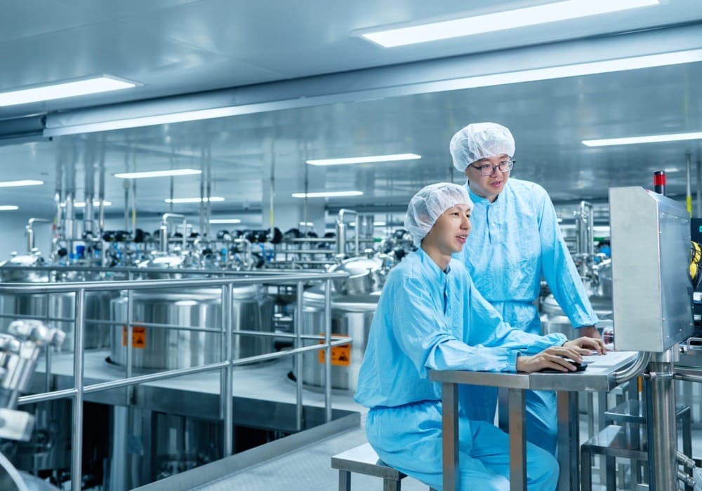 Two technicians in blue uniforms and hairnets operating a computer in a large sterile industrial facility.