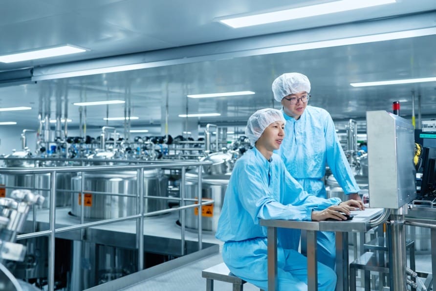 Two technicians in blue uniforms and hairnets operating a computer in a large sterile industrial facility.