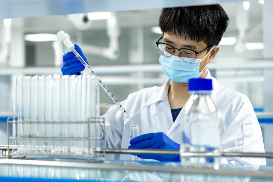 Scientist in a lab coat and mask using a pipette to handle liquid samples in a laboratory setting with test tubes nearby